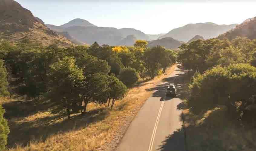 A car drives down a two-lane road surrounded by trees. Dry mountains rise in the distance.